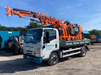 Camion blanc avec une grue orange montée sur plateau, stationné sur un sol en gravier avec des véhicules et conteneurs en arrière-plan.