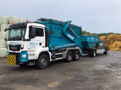 Excavatrice jaune chargée de débris métalliques dans une benne verte sur un chantier en terrain accidenté.