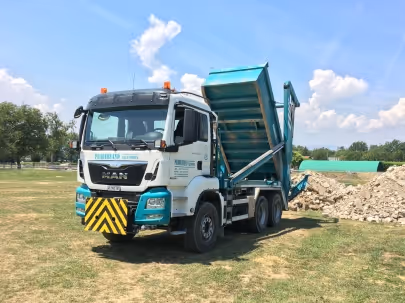 Camion benne blanc et bleu avec lit de chargement incliné déversant des gravats sur un terrain herbeux sous ciel bleu.