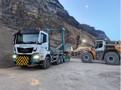 Camion blanc avec un équipement de levage et un chargeur sur un chantier minier au pied d'une montagne rocheuse au crépuscule.