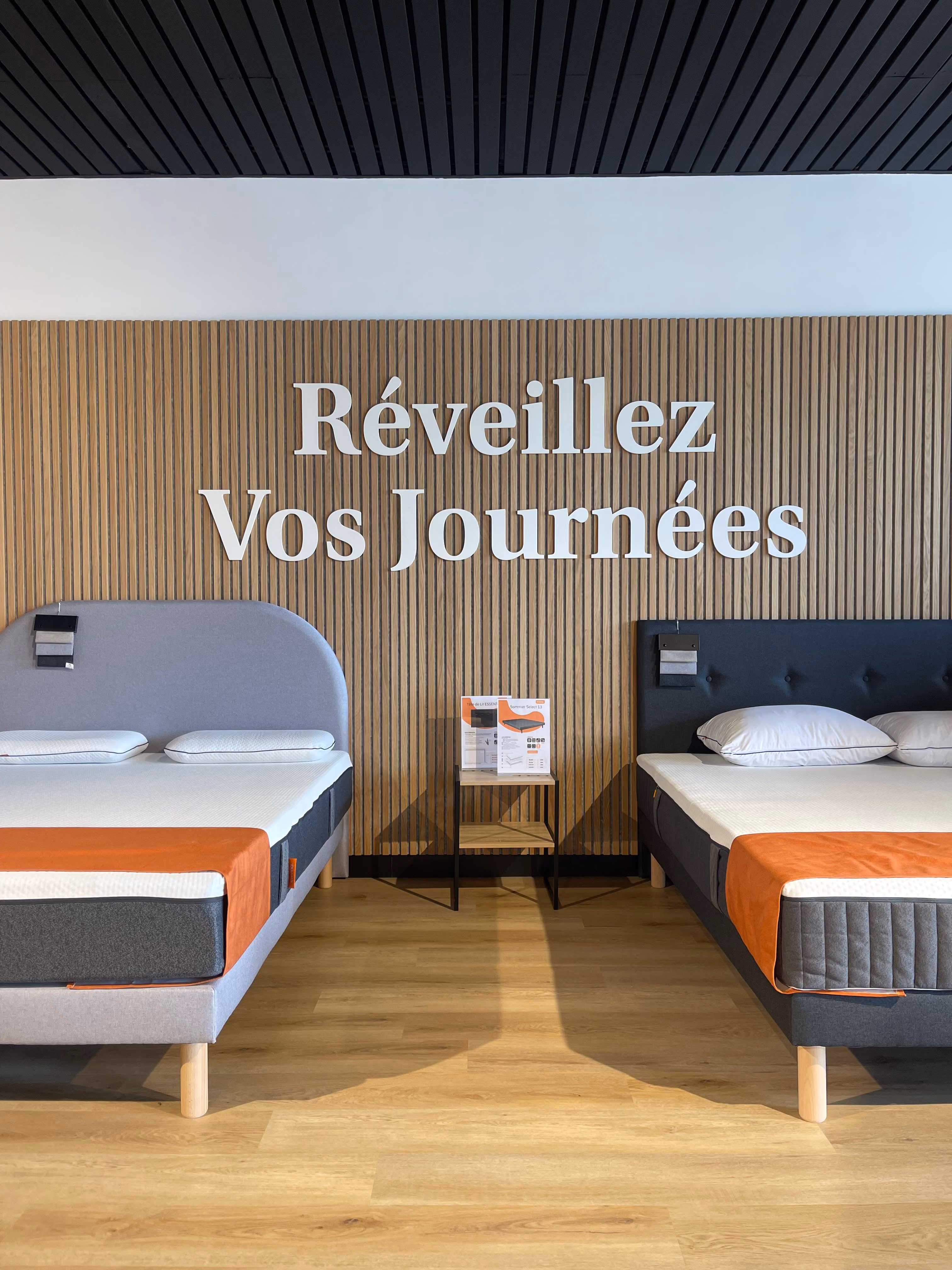 Two beds with pillows and orange runners in a showroom with wooden slat wall and 'Réveillez Vos Journées' text.