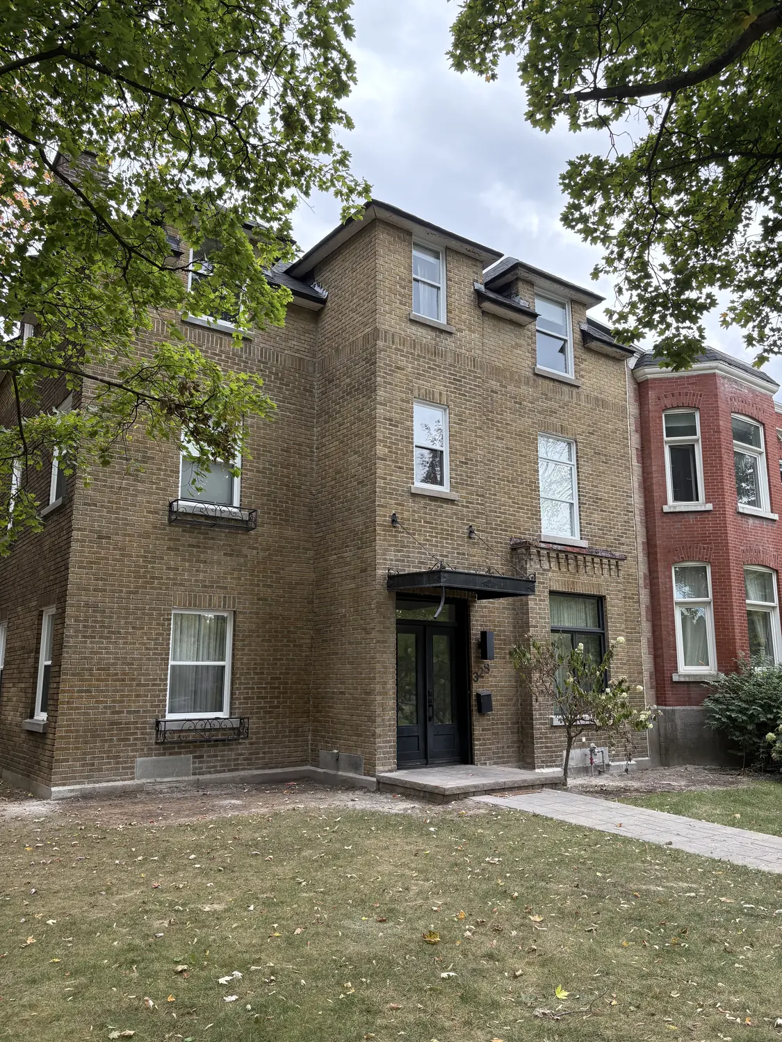 Bâtiment résidentiel en brique beige de trois étages avec une entrée noire, entouré d'arbres et d'une pelouse.