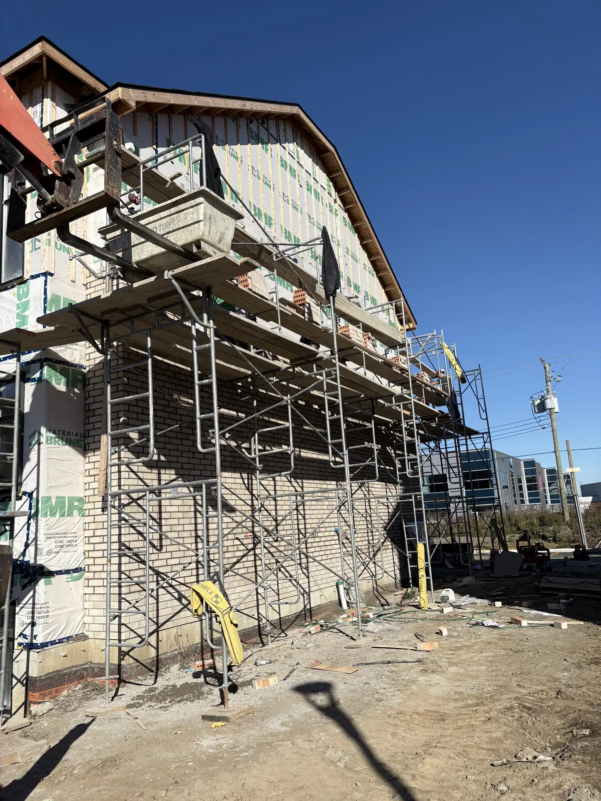 Chantier de construction avec échafaudage métallique devant un mur en briques partiellement revêtu d'une membrane étanche sous un ciel bleu clair.