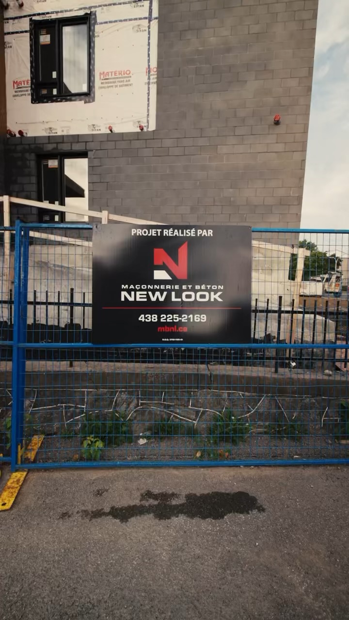 Chantier avec une clôture bleue et une affiche noire indiquant 'Projet réalisé par Maçonnerie et Béton New Look' avec un logo rouge, un numéro de téléphone et un site web.