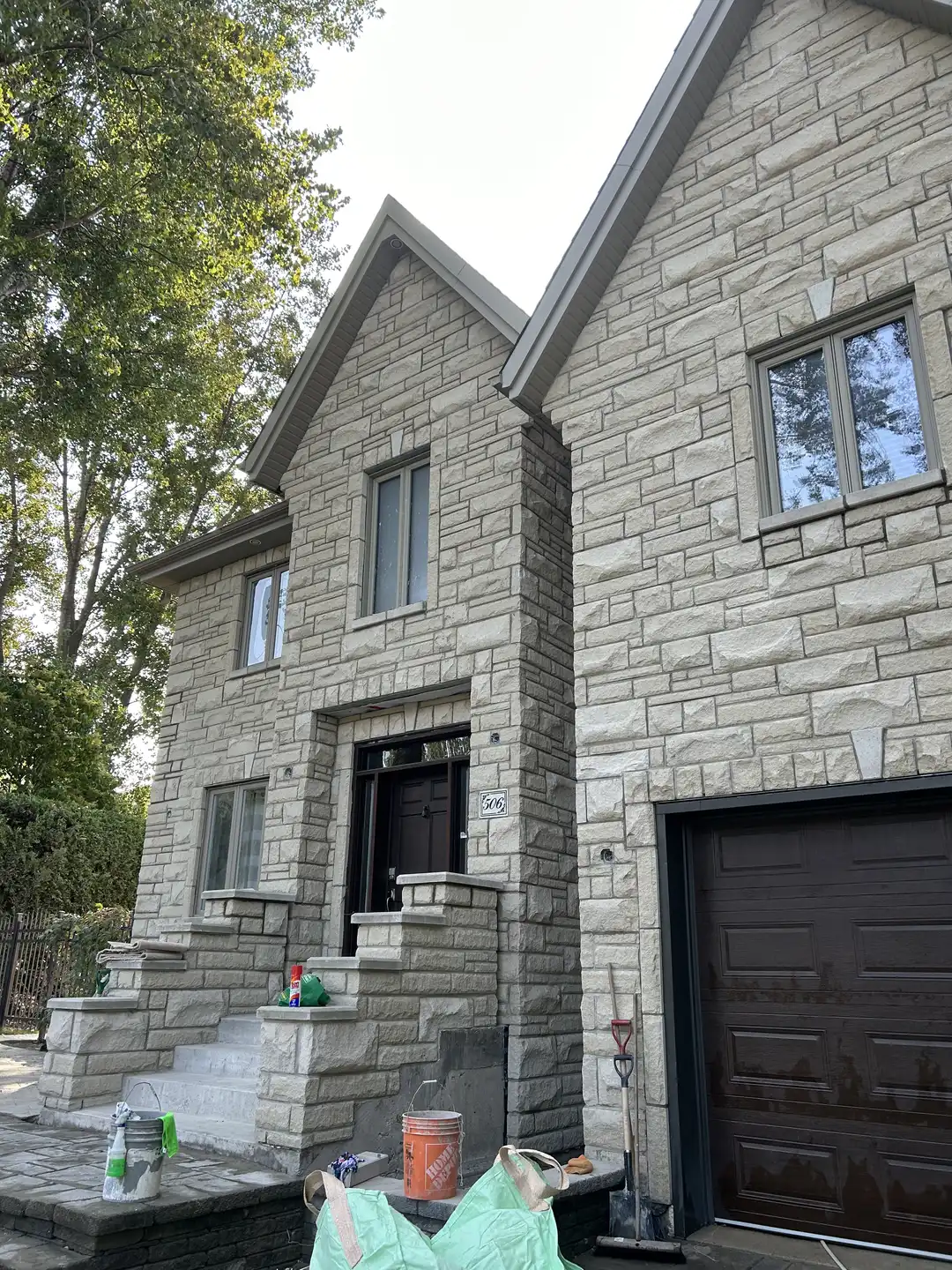Maison en pierre beige avec escalier à l'entrée, porte noire, garage brun et outils de jardin posés devant.