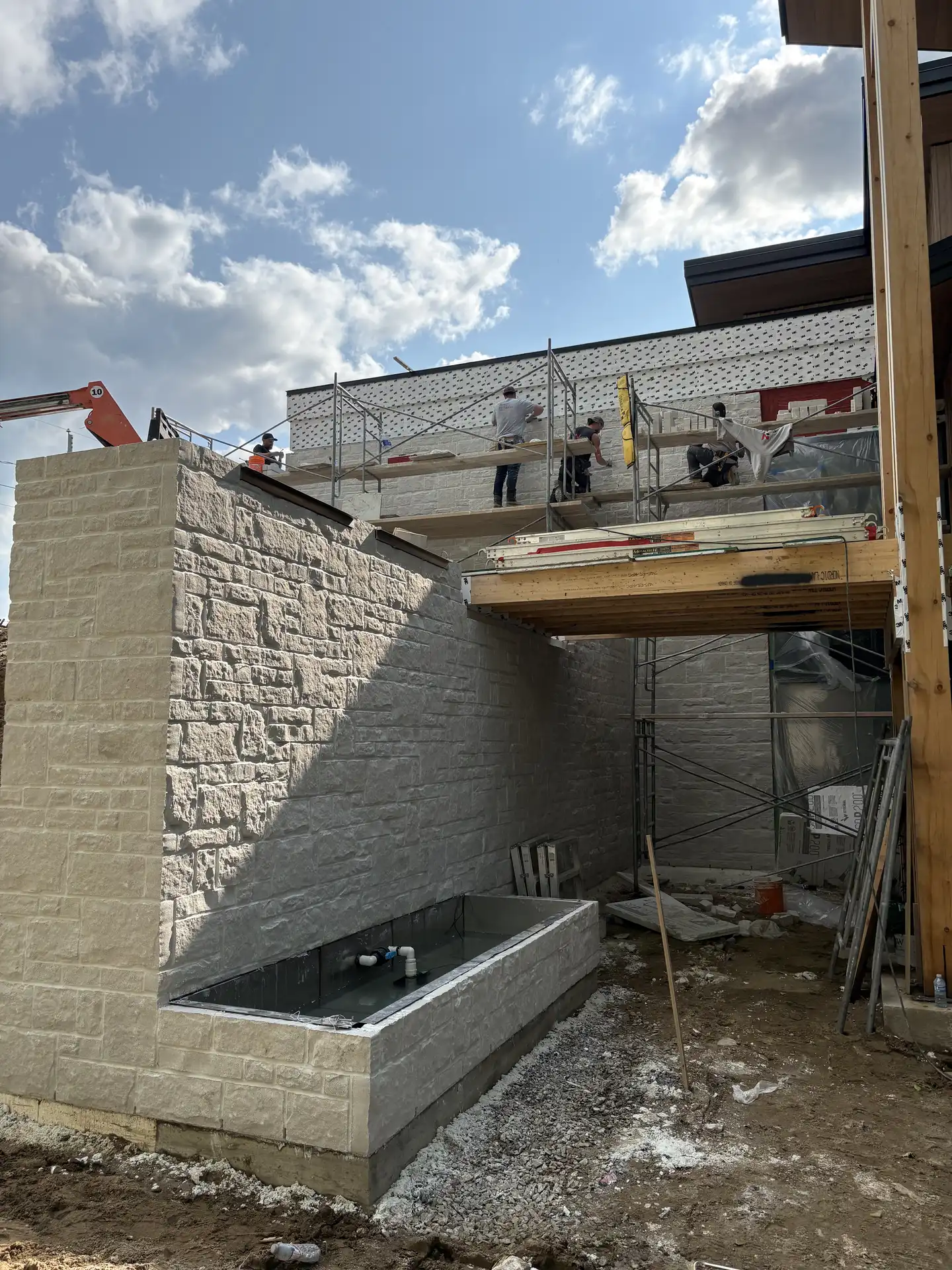Travailleurs sur échafaudage construisant un mur en pierre avec un bassin rectangulaire vide au premier plan.