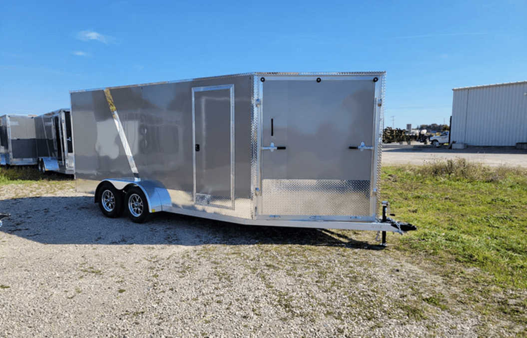 Silver enclosed trailer with two axles parked on gravel near grass and buildings under clear sky.