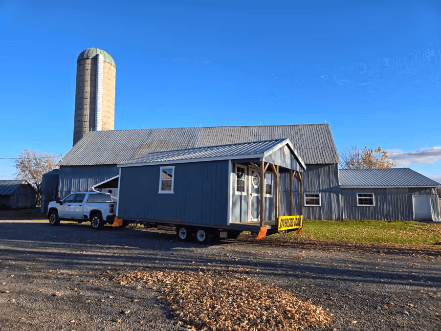 Blue tiny house on wheels being towed by white pickup truck in front of a barn and silo.