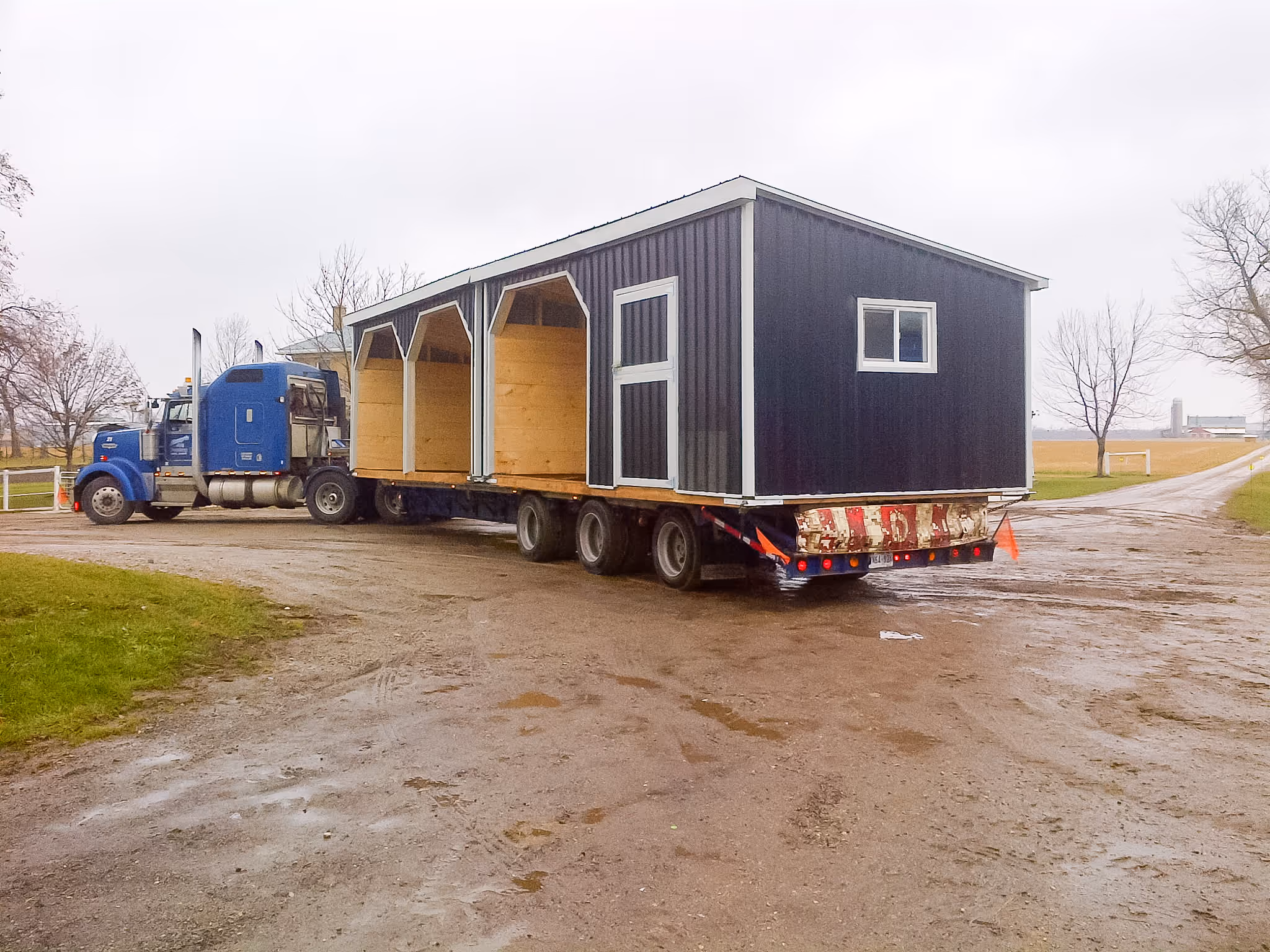 Large blue truck hauling a trailer with a black and wood tiny house on a rural dirt road.