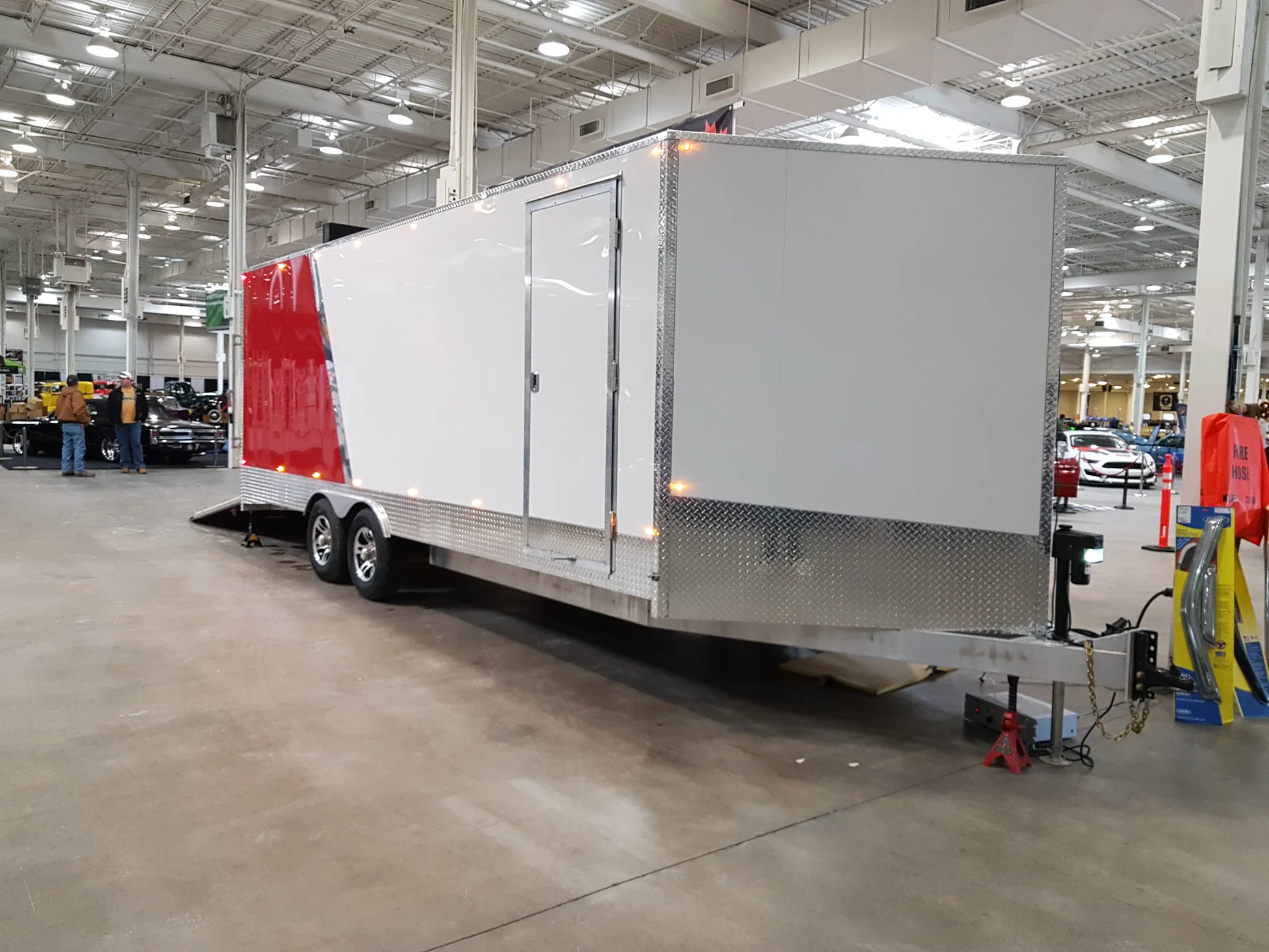 Large enclosed car hauler trailer with white and red panels parked indoors at a car show.