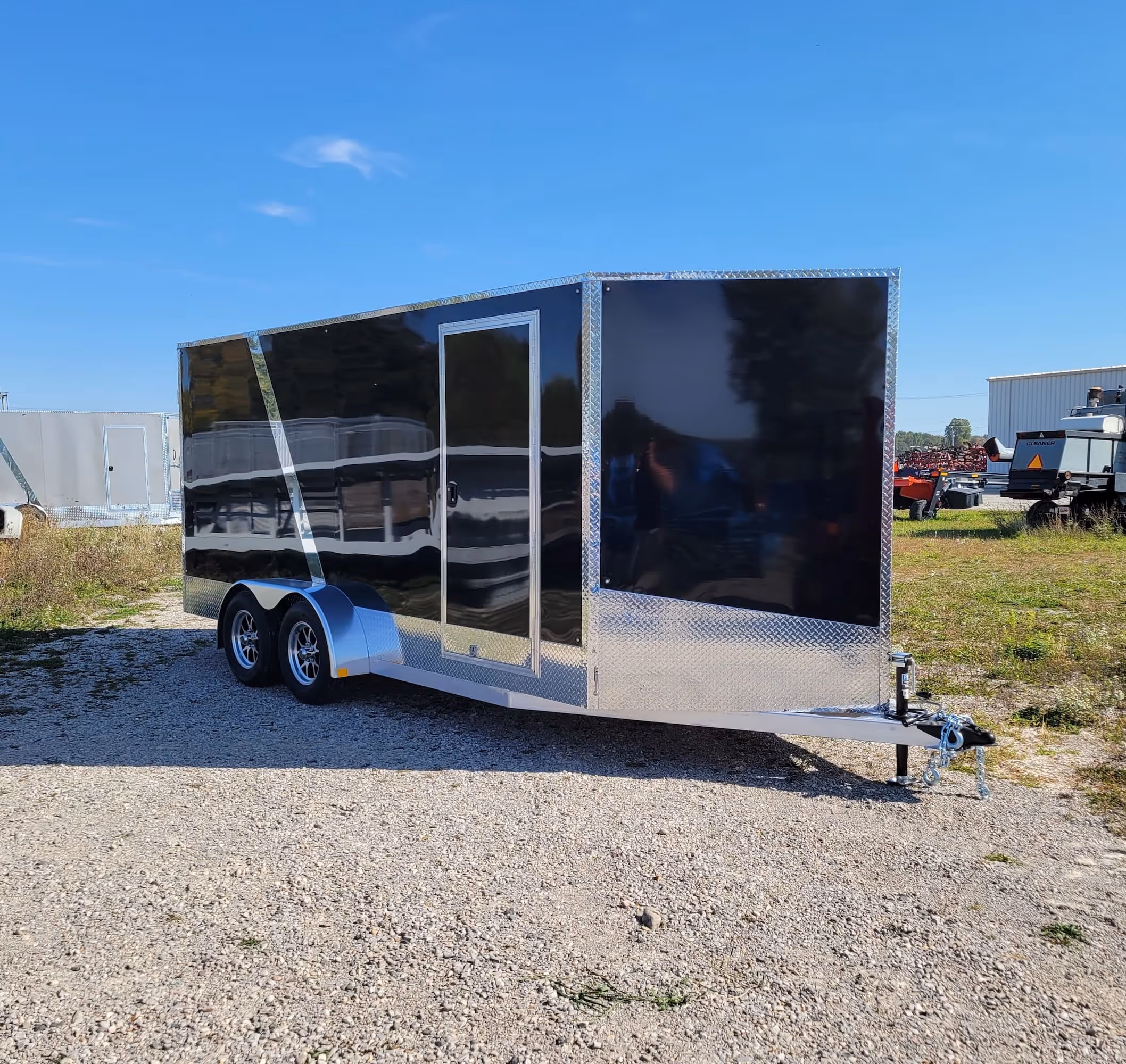 Black enclosed cargo trailer with silver trim and dual axles parked on gravel under blue sky.