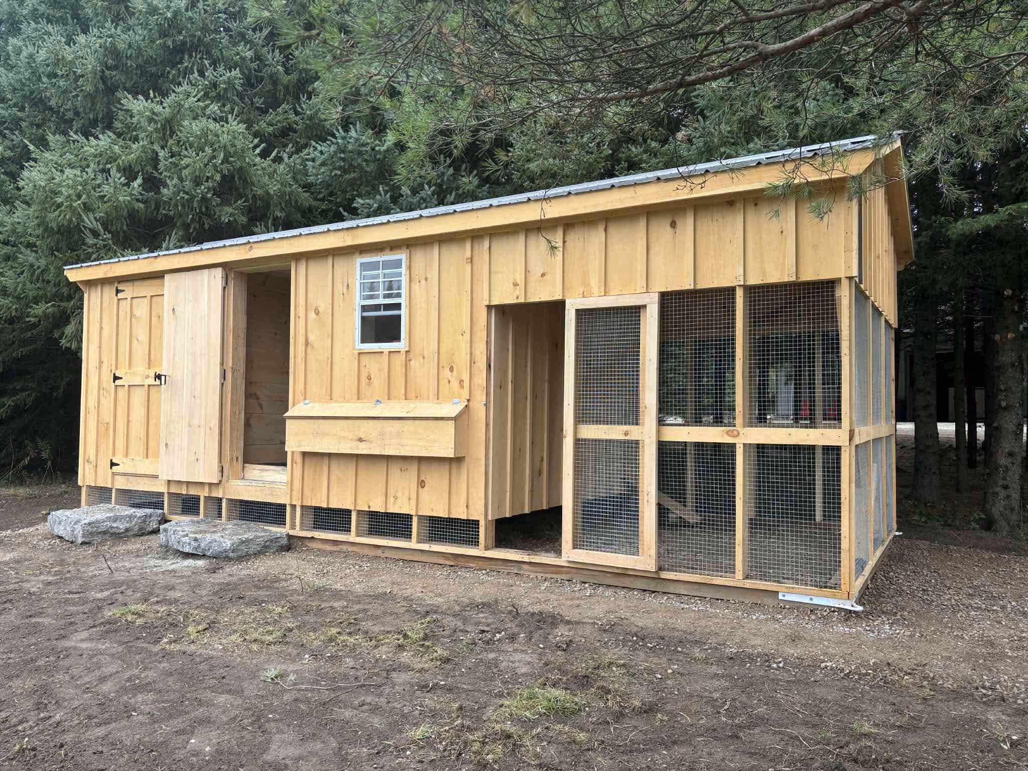 Wooden chicken coop with open doors, a window, and attached wire mesh run near trees.