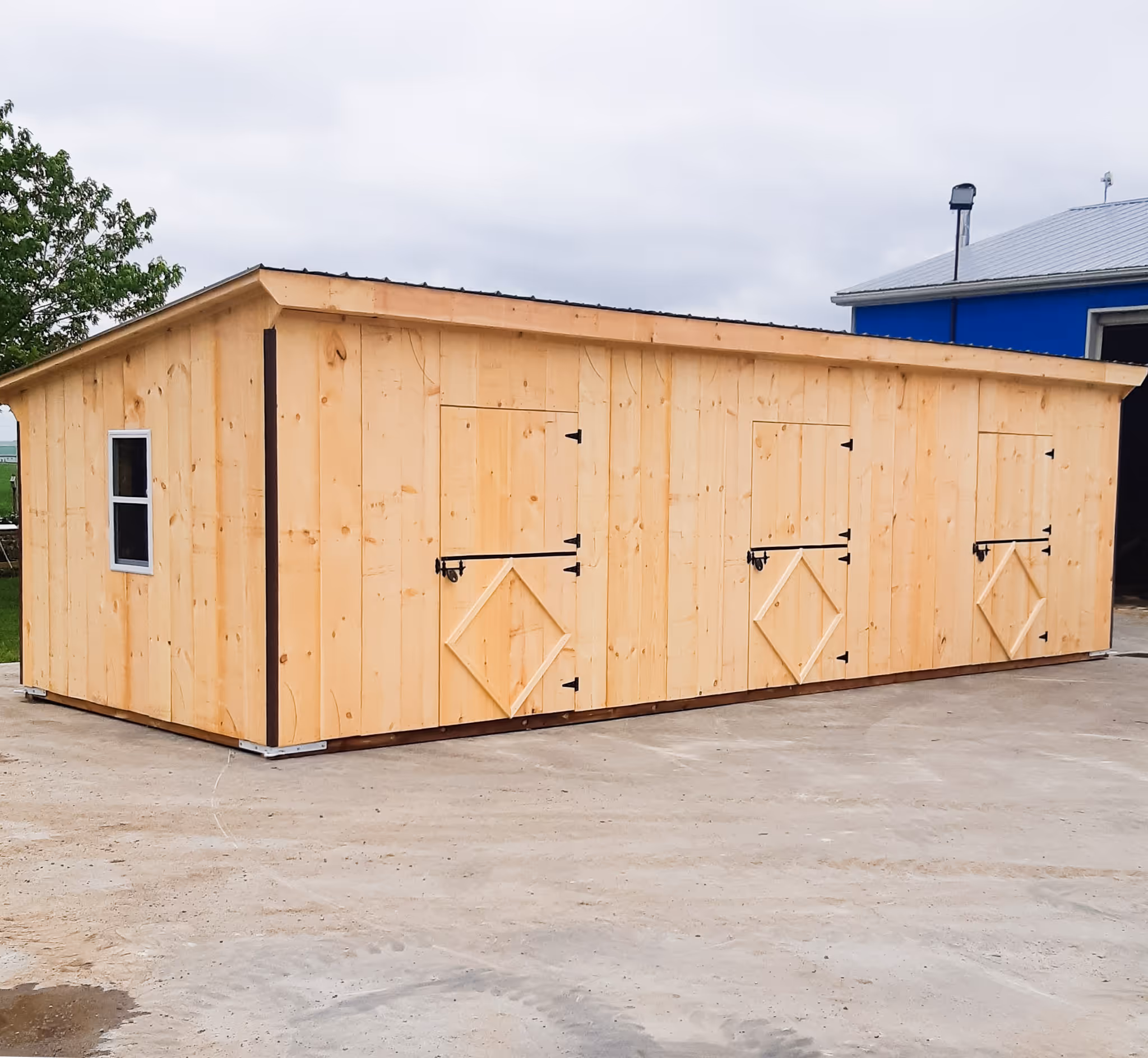 Long wooden shed with three doors, a side window, and a slanted roof on a concrete surface.