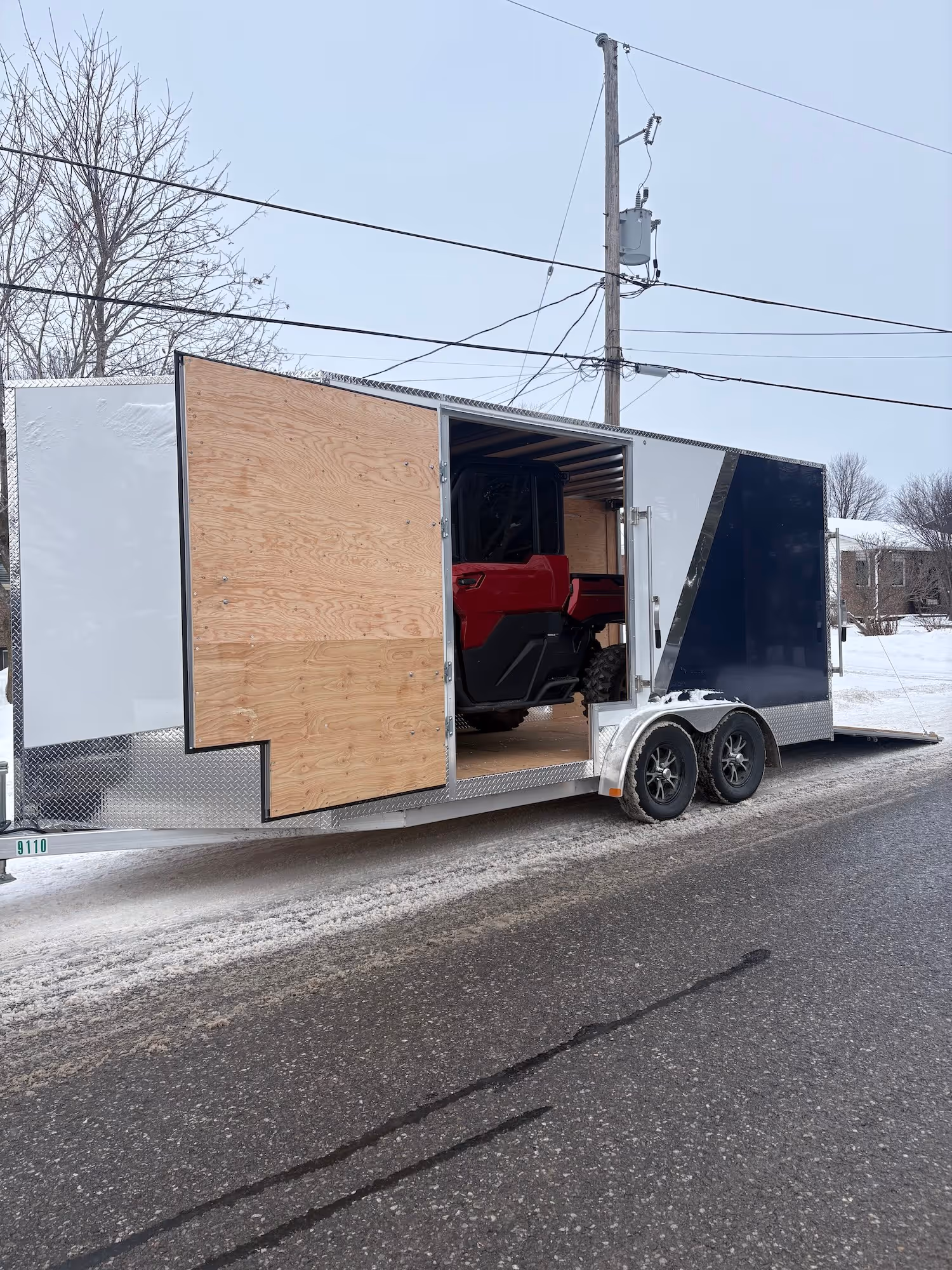 White and blue enclosed trailer parked on snowy road carrying a red off-road vehicle.