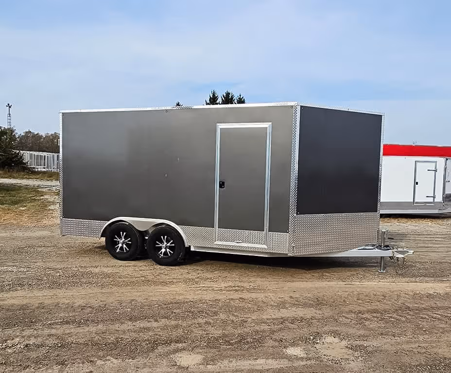 Gray enclosed trailer with dual axles and a side door parked on a dirt surface under a clear sky.