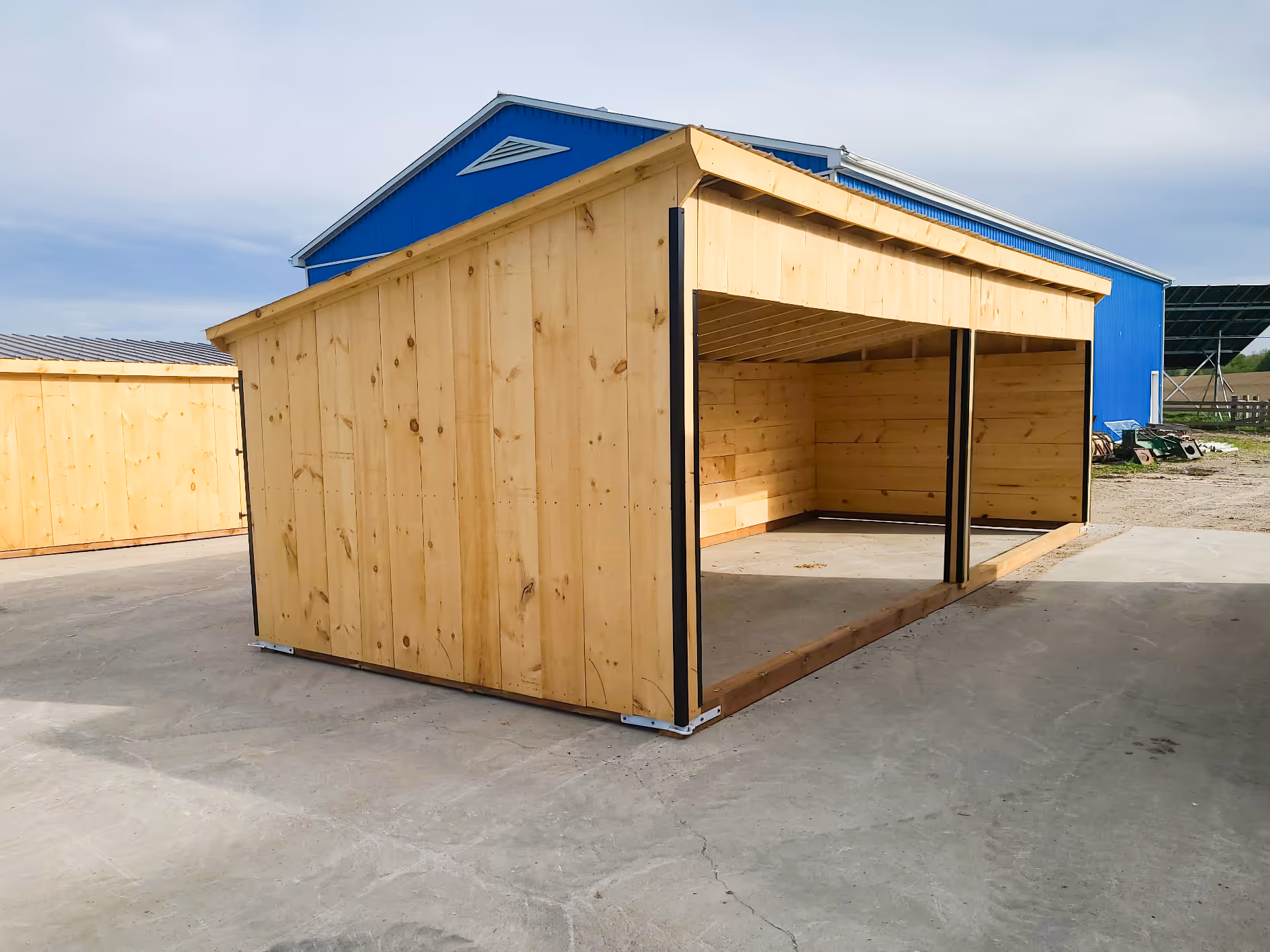 Large wooden open-sided shelter structure on concrete near blue building under clear sky.