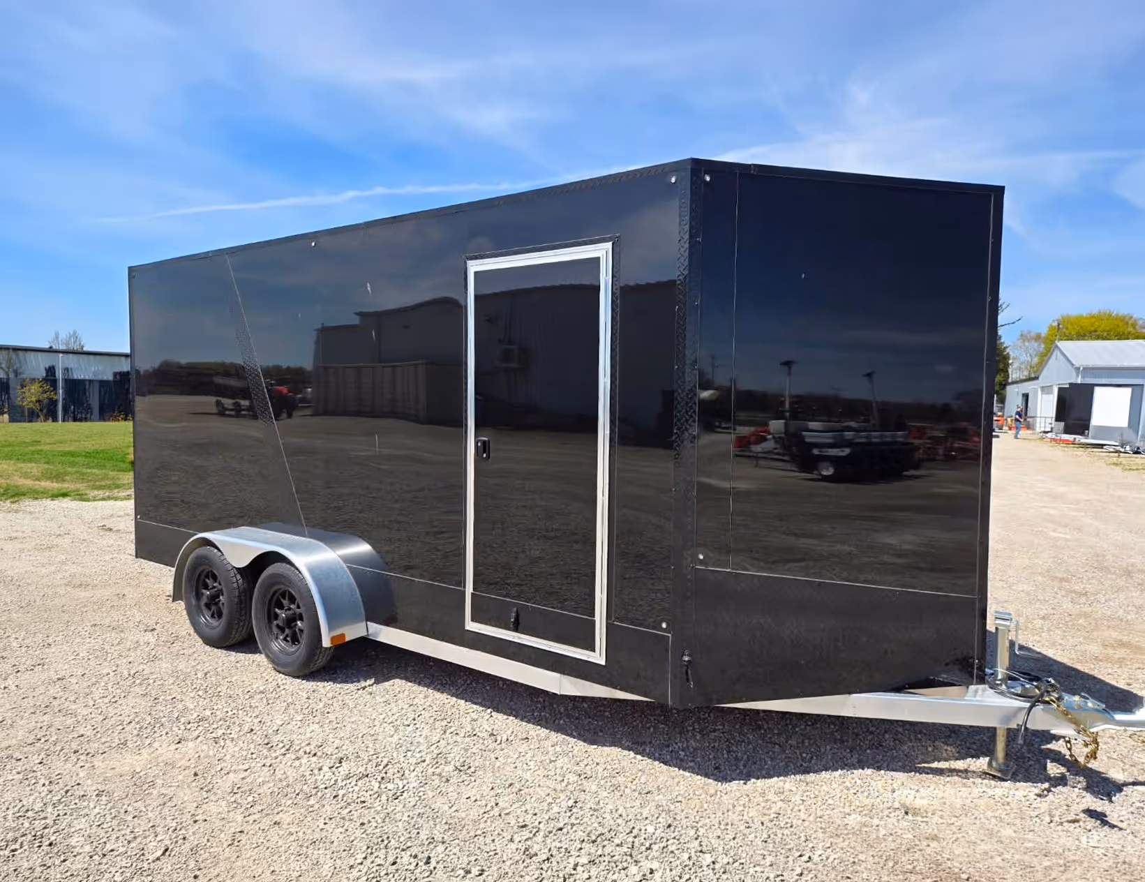Black enclosed dual-axle trailer with a side door parked on gravel under a blue sky.