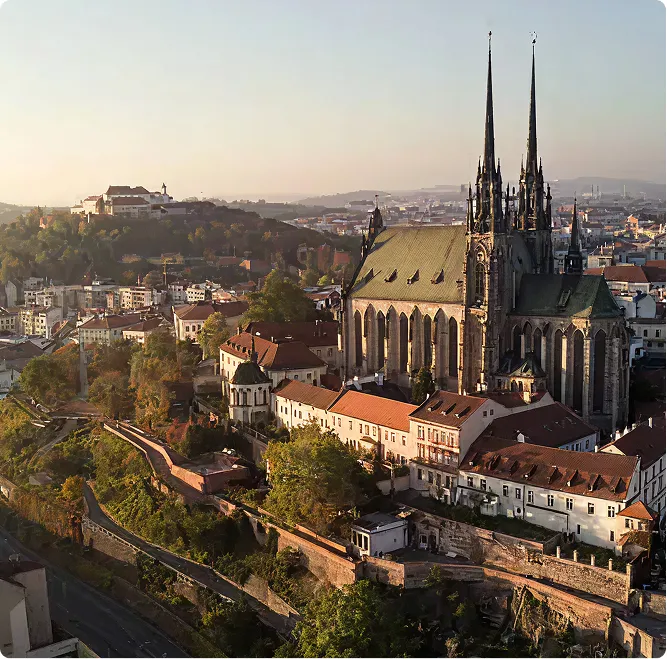 Pohled na historickou katedrálu svatého Petra a Pavla v Brně s okolními budovami a zalesněným kopcem v pozadí za slunečného dne.