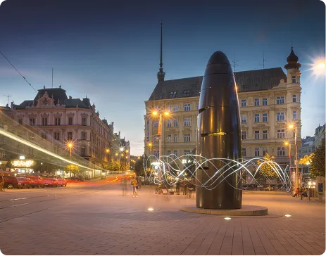 Noční městské náměstí s moderní černou sochou a historickými budovami v pozadí osvětlenými pouličními lampami.
