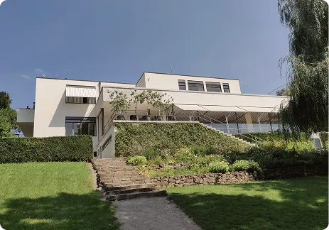 Modern white building with a terrace, stairs, and a green lawn under a clear sky.