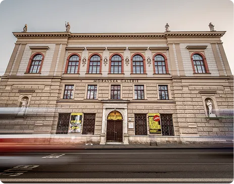 Historická budova Moravské galerie s okny a sochami za pohybujícími se auty.