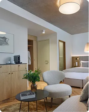 Modern hotel room with bed, round chair, wooden cabinet, coffee machine, and a vase with greenery.