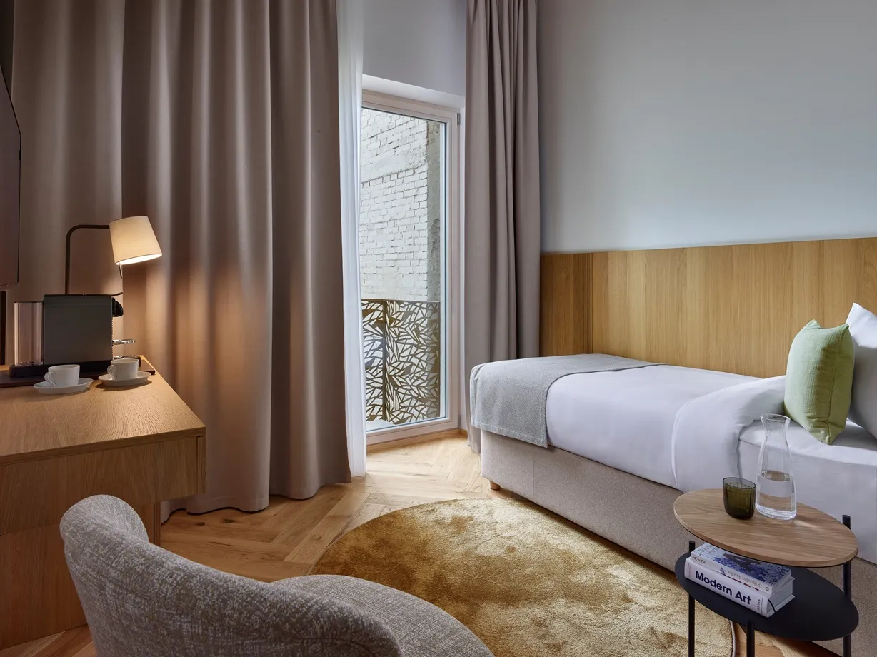 Modern hotel room with single bed, wood furnishings, round rug, and a small balcony door.