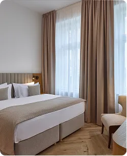 Modern bedroom with beige bed, white pillows, tall windows, and beige curtains.