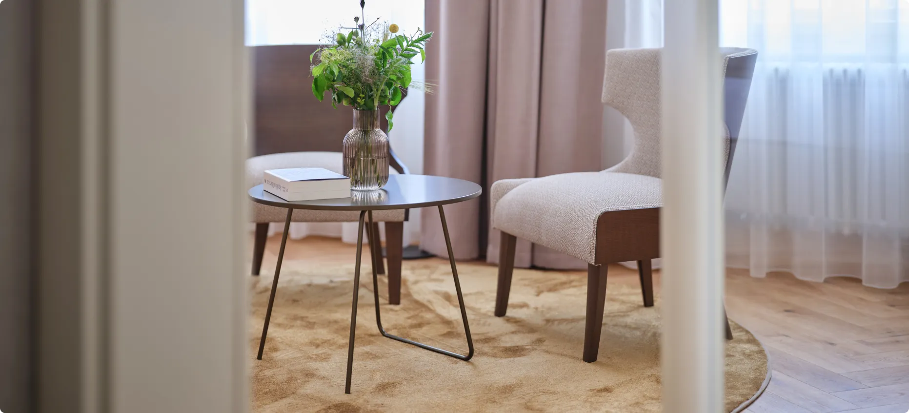 Modern cozy seating area with round table, flower vase, book, two beige chairs, and soft rug.