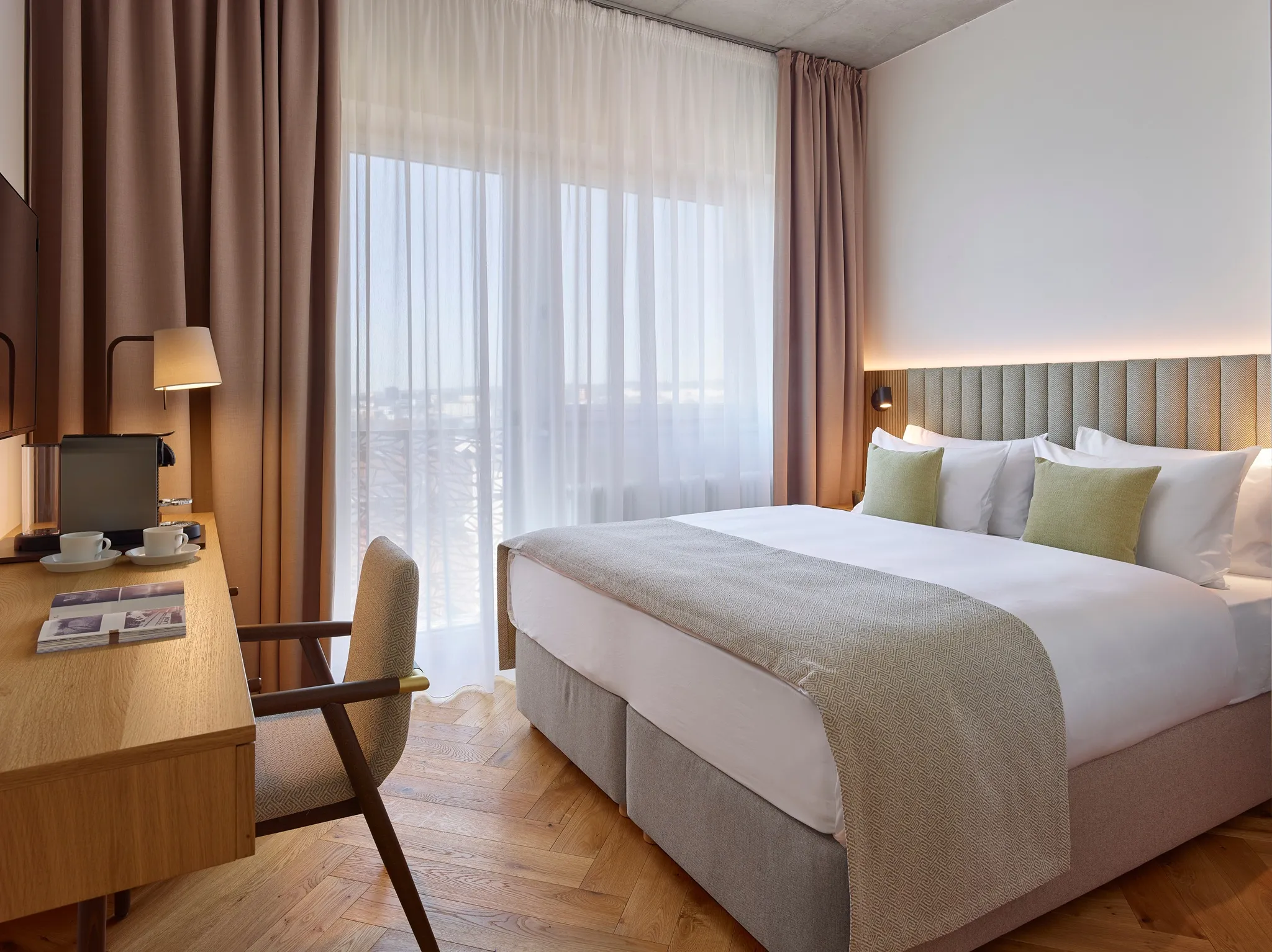Modern hotel room with large bed, beige and green pillows, wooden desk, chair, and sheer curtains.