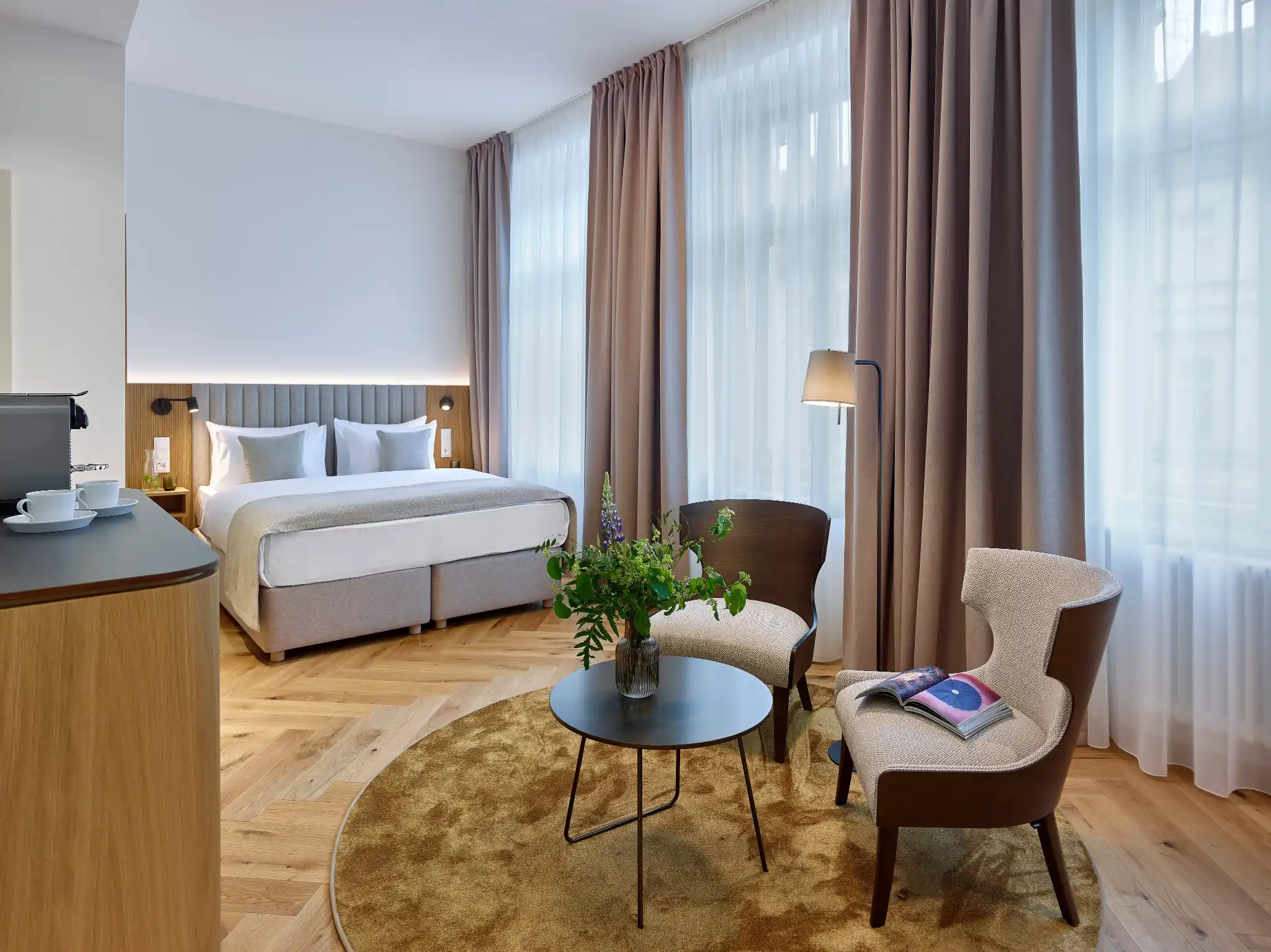 Modern hotel room with double bed, two armchairs around a round table with flowers, and large windows with curtains.