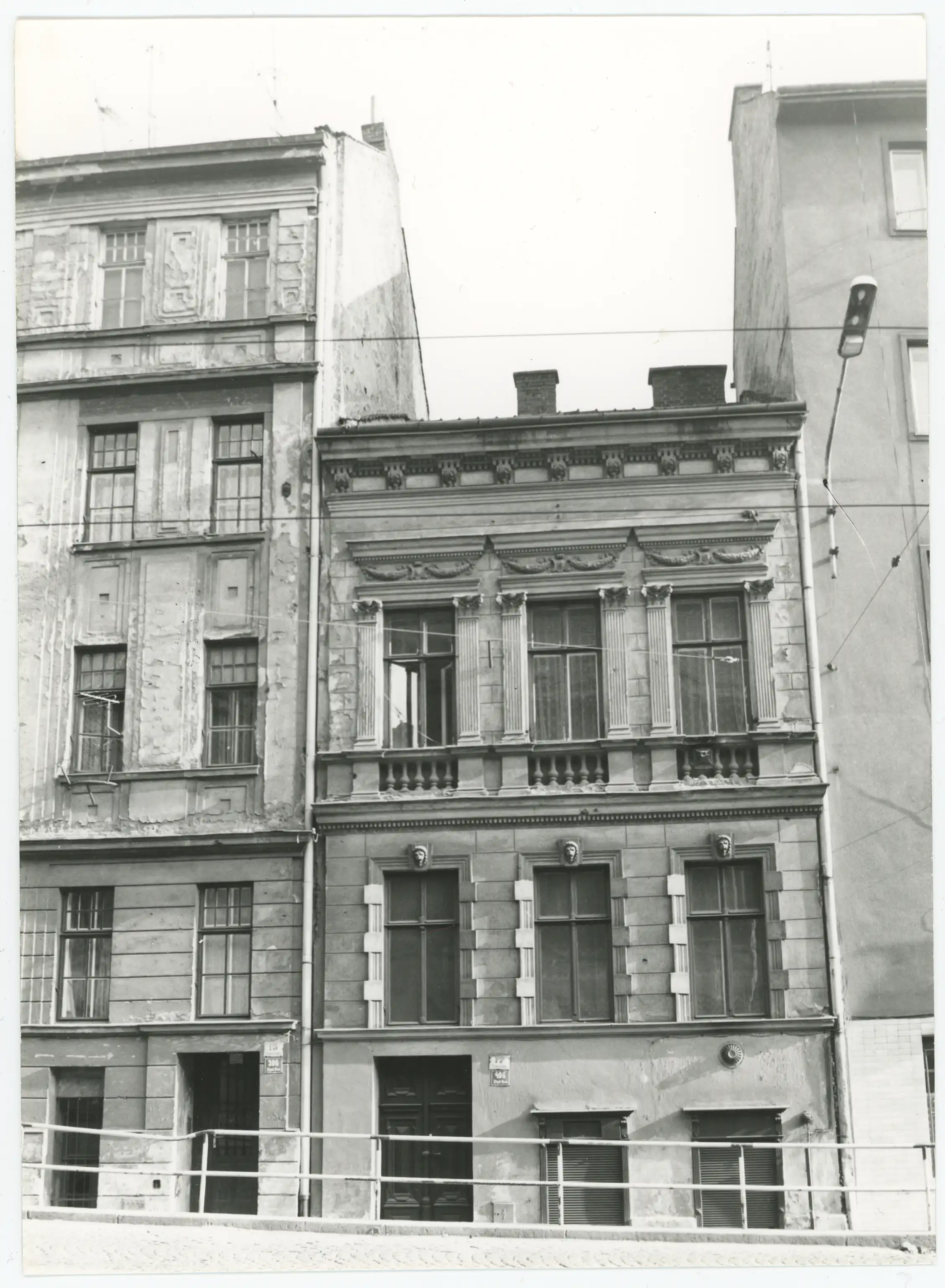 Černobílá fotografie starých zdobených městských domů se dvěma vchody a oplocením v popředí.