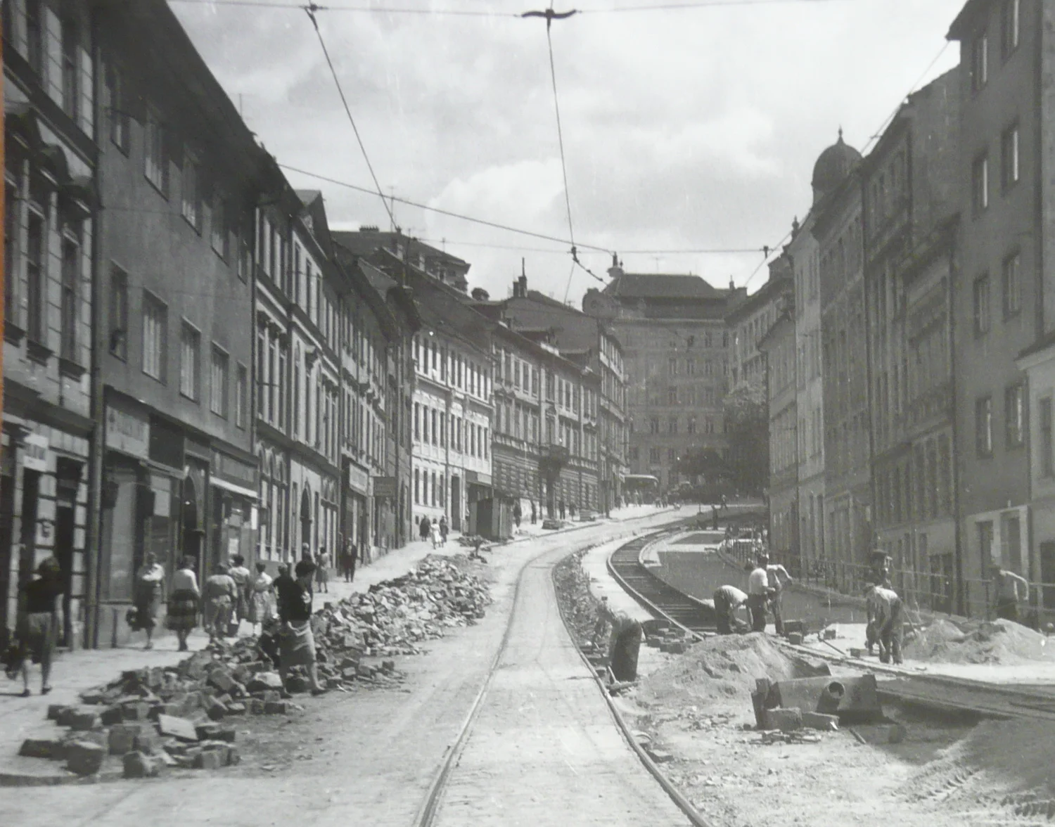 Černobílá fotografie ulice ve městě s opravami kolejnic a chodníku, lidé chodí po stranách.