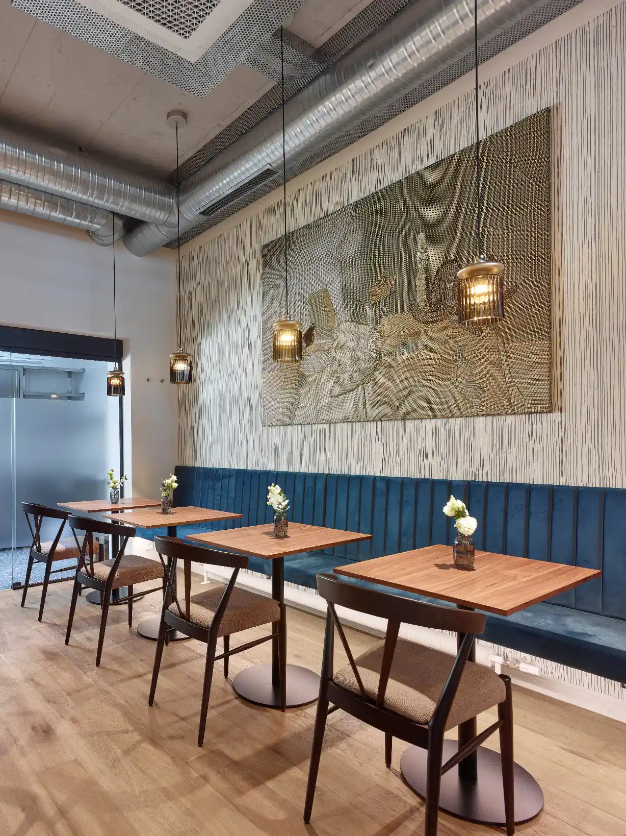 Modern cafe interior with wooden tables, dark chairs, blue bench, hanging lights, and wall art.