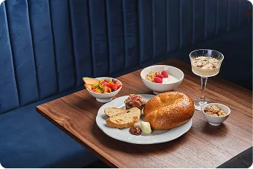 Snídaně na dřevěném stole: bagel, máslo, dýňová semínka, ovoce a jogurt v miskách a sklenici.