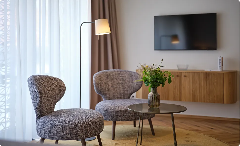 Dvě šedé křesla u stolu s vázičkou květin, stojací lampa a televizor na stěně v obýváku.