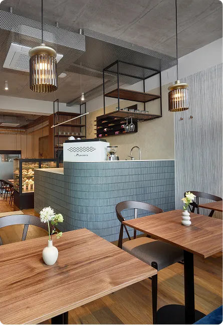 Modern café interior with wooden tables, small flower vases, blue tiled counter, and hanging pendant lights.