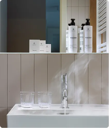 Two clear glasses on bathroom sink with running water and white skincare bottles on shelf.