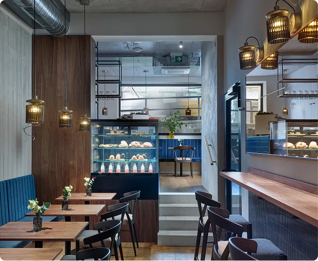 Modern café interior with wooden tables, black chairs, blue bench, pendant lights, and display case with pastries.