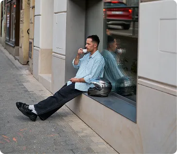Muž sedící na parapetu u okna na ulici, pije kávu, má vedle sebe motocyklovou helmu.
