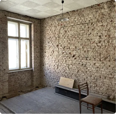 Prázdná místnost s holou cihlovou zdí, oknem a dřevěnou židlí na prachu podlahy.