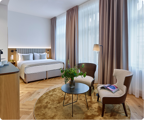 Modern hotel room with double bed, round rug, two chairs, table with flowers, and large windows with curtains.