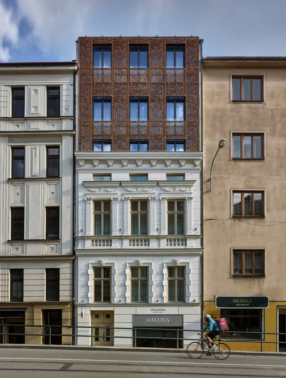 Modern white building with decorative upper facade and cyclist passing in front on a city street.