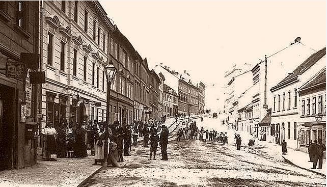 Černobílý historický snímek ulice s chodci a budovami na obou stranách.