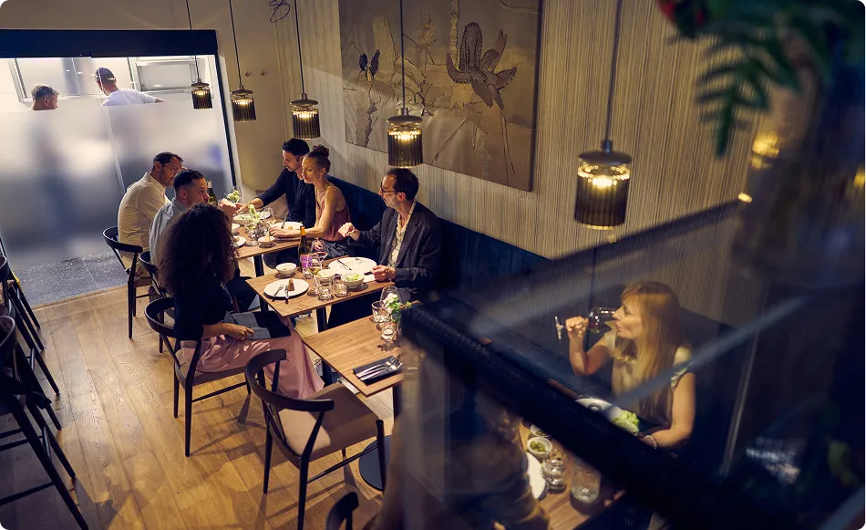 Skupina lidí sedící u stolu v restauraci při konverzaci a jídle ve večerním osvětlení.