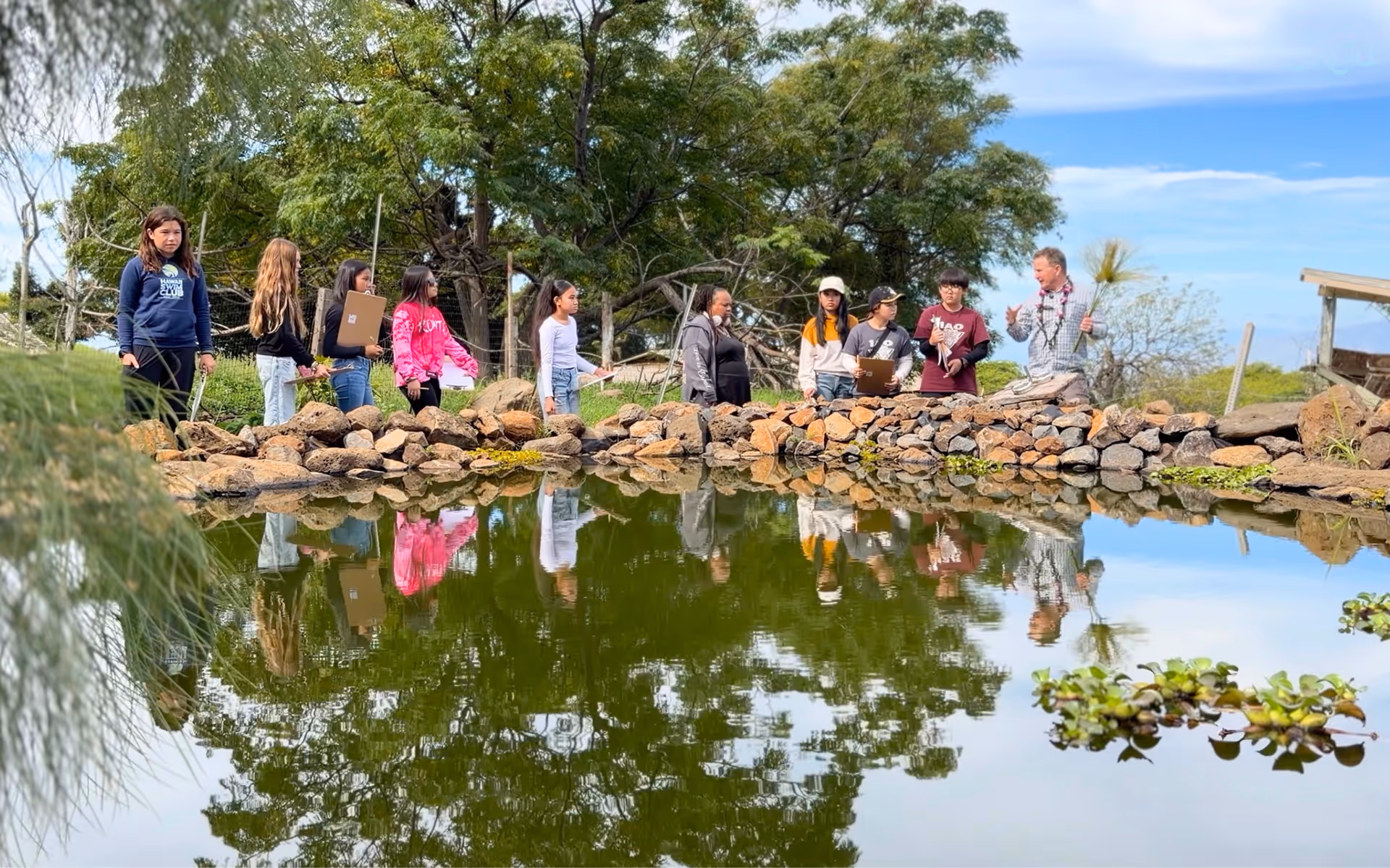 Group of students and a teacher standing by a pond with rocks, some holding clipboards, engaged in outdoor learning.