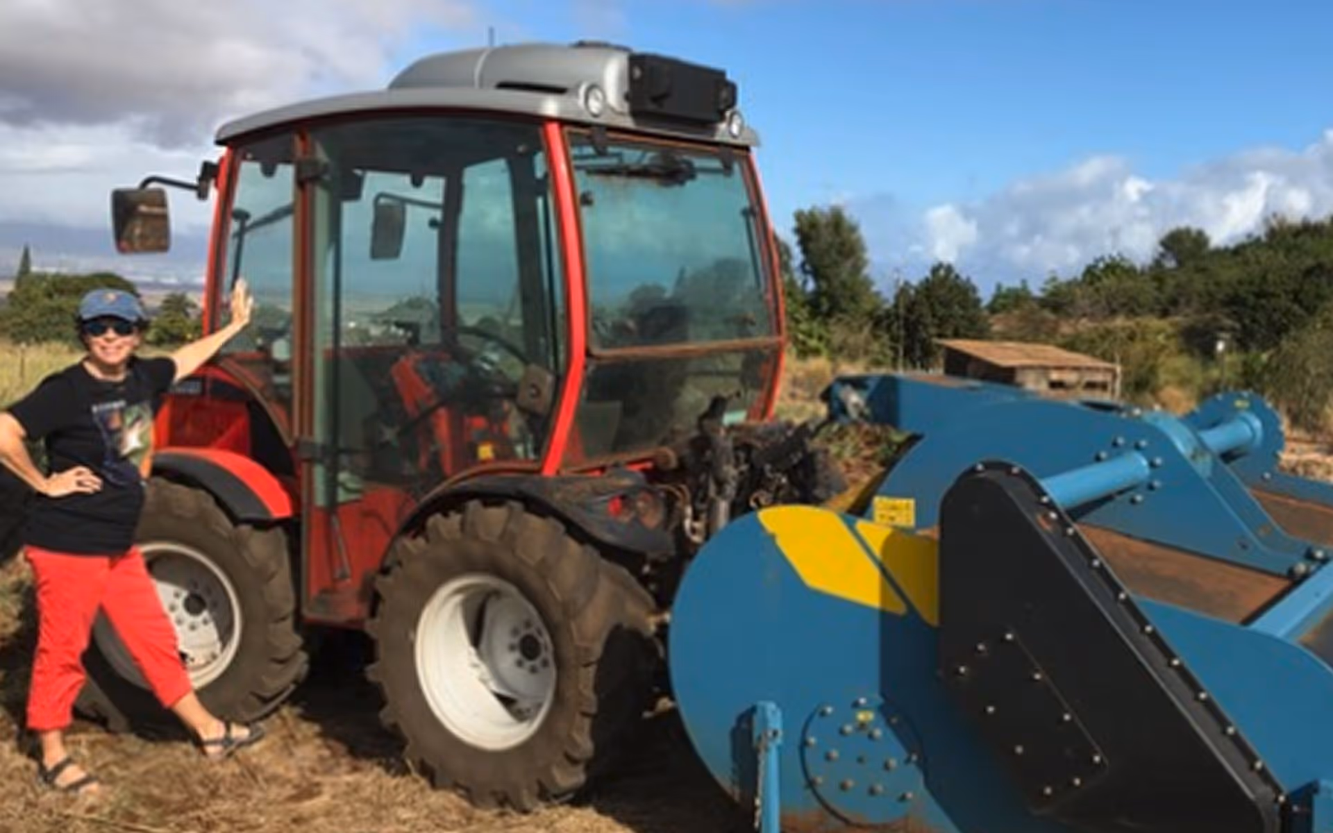 Person in a blue cap and red pants standing next to a red tractor with large rear tires and a blue agricultural attachment on a sunny day.