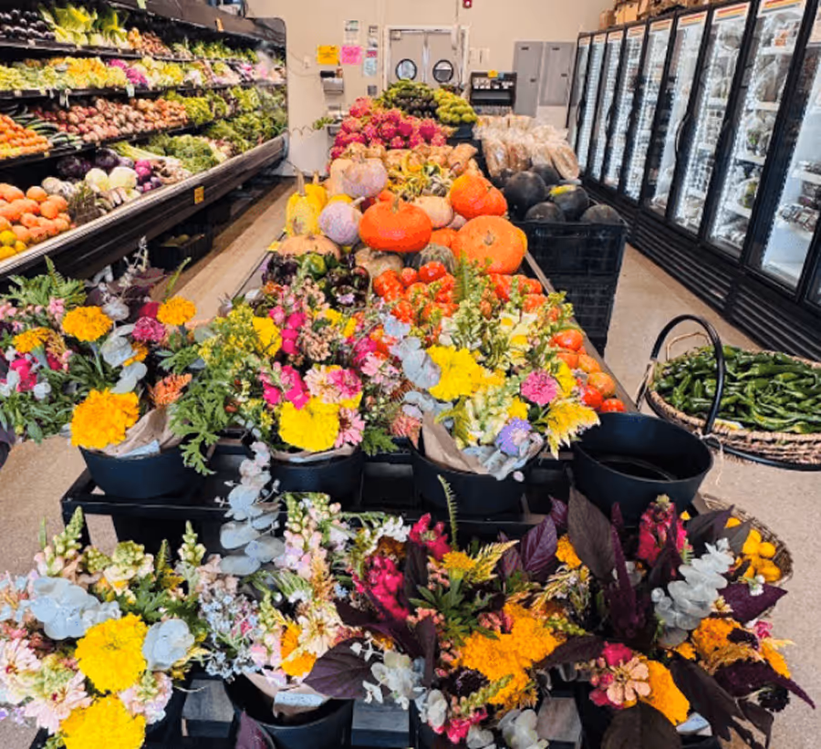 Indoor market with colorful flower bouquets in the foreground and fresh vegetables and pumpkins displayed on tables and shelves in the background.