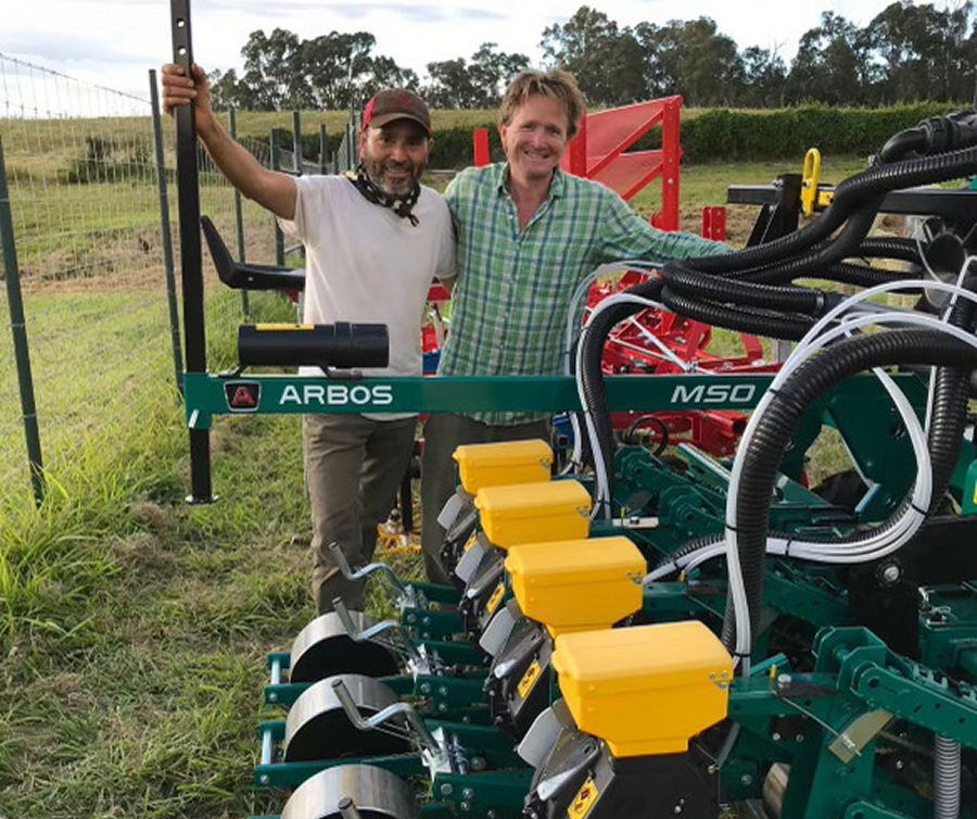 Two men standing outdoors by an Arbos M50 agricultural machine in a grassy field.