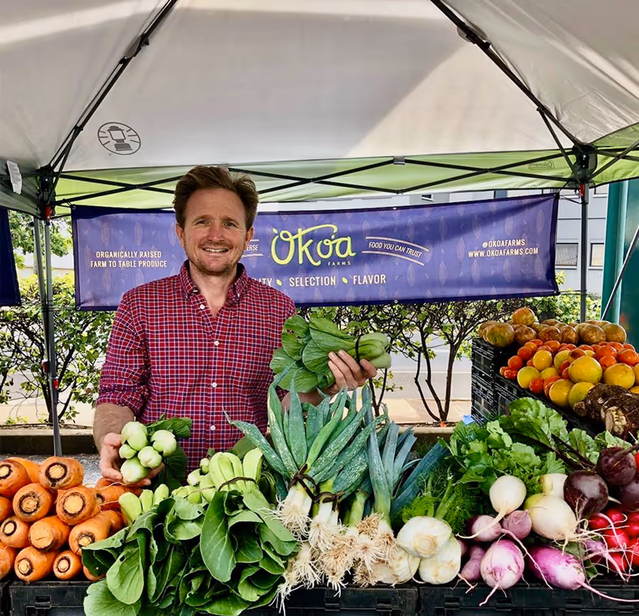 Man in red plaid shirt standing behind farmer's market stall with fresh vegetables including carrots, bok choy, leeks, turnips, and tomatoes under a canopy.